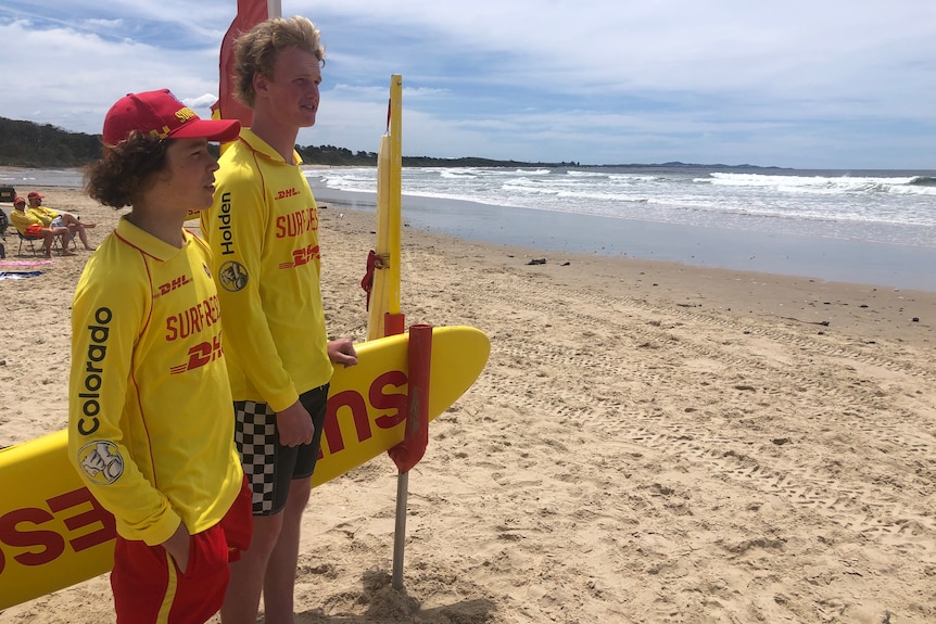 Two teenage boys wearing lifesaver uniforms look over a beach. 