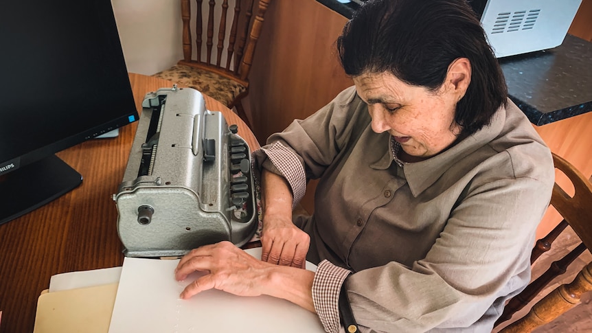 A woman sits at a kitchen table with her hands on a Braille writer