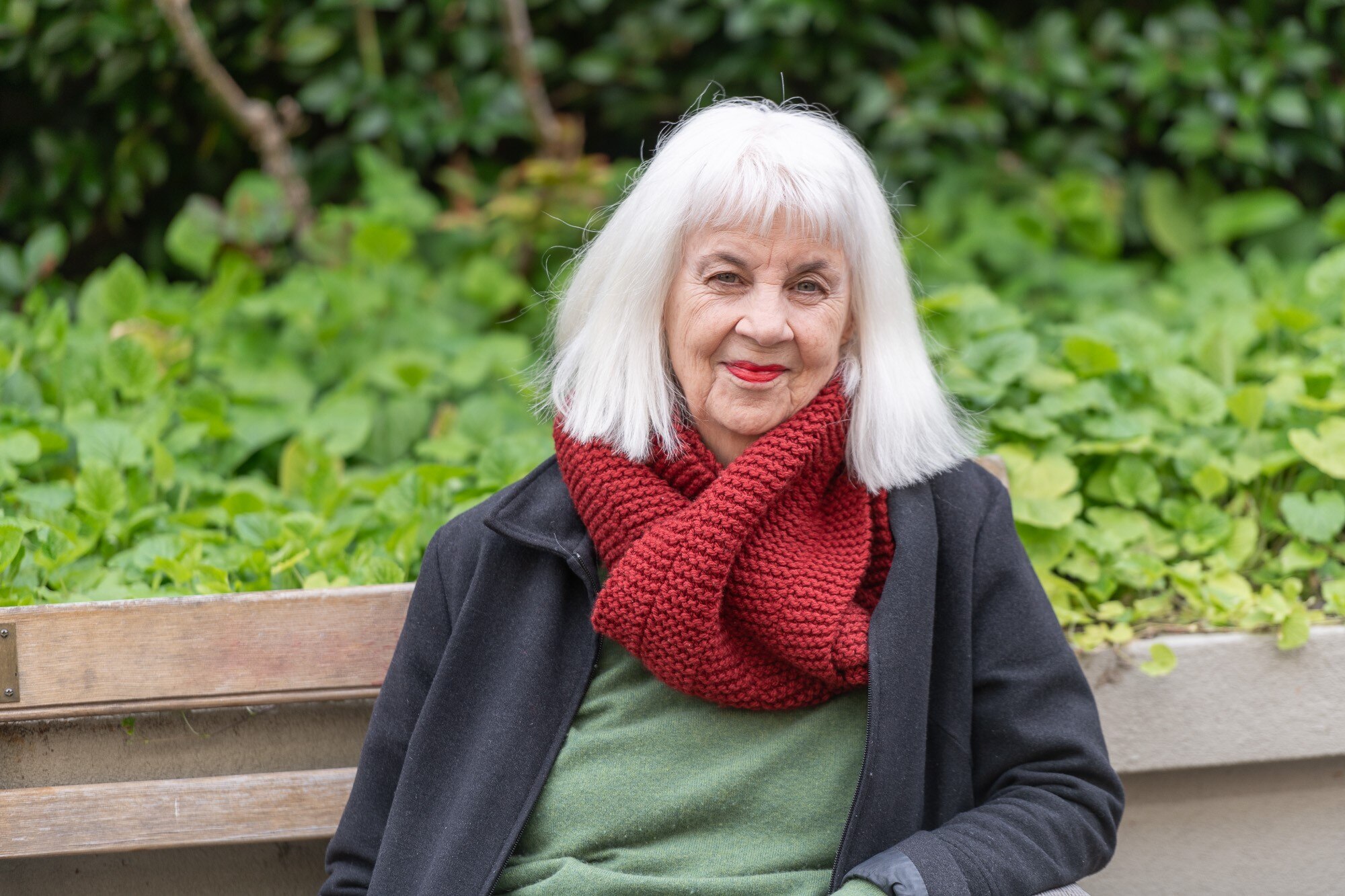 A white woman in her 70s with white shoulder length hair and wearing a red scarf sits on a bench in a garden