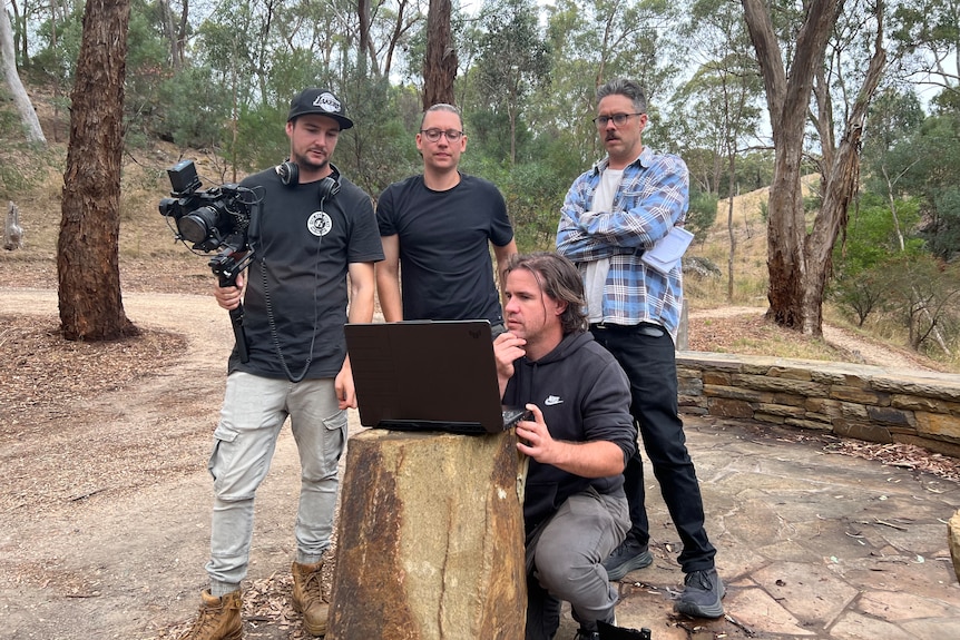A group of men stand over a computer in the middle of the bush watching a laptop screen