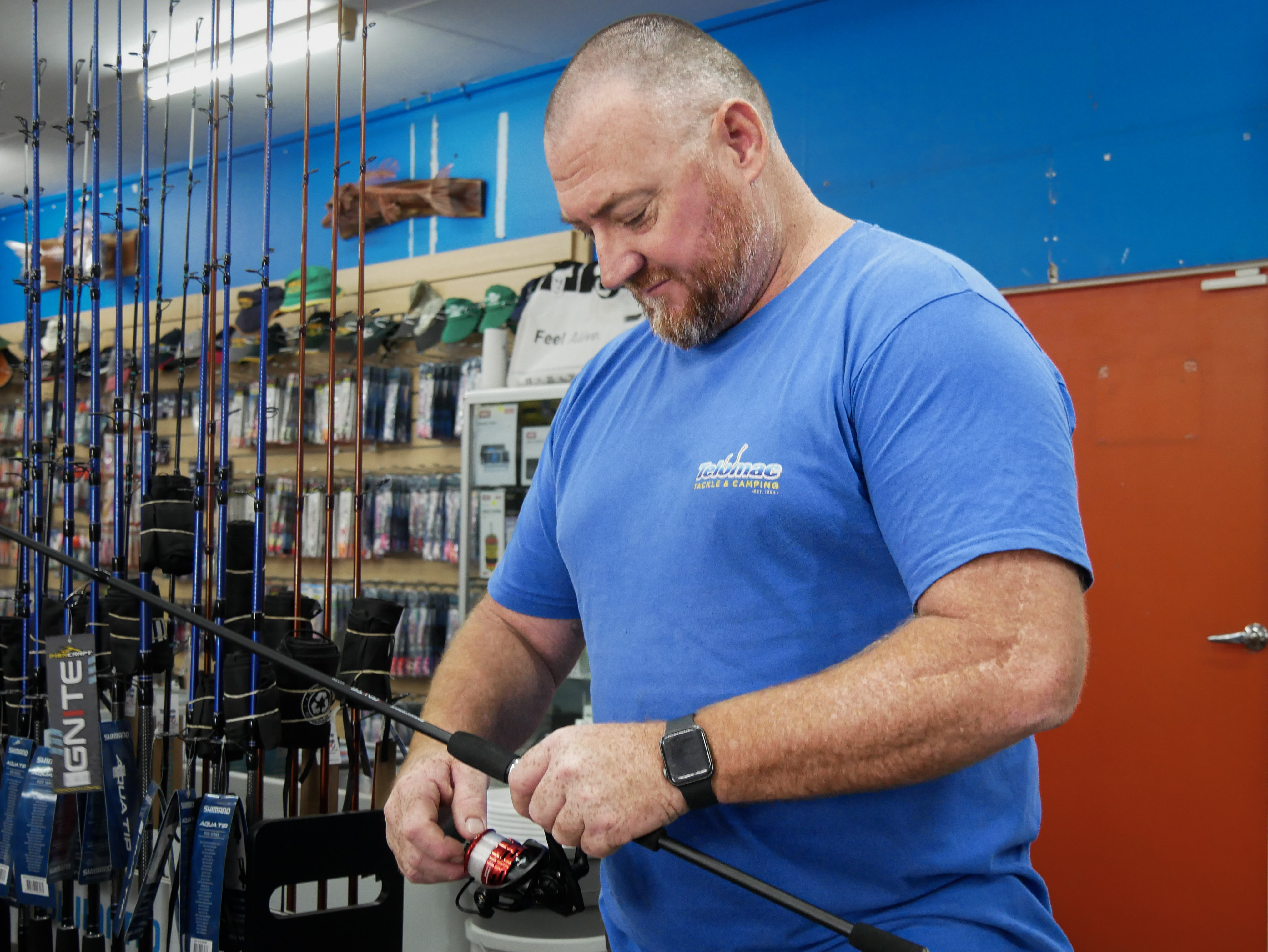 Um homem de camisa azul verifica o carretel de uma vara de pescar no balcão de uma loja de iscas e equipamentos.