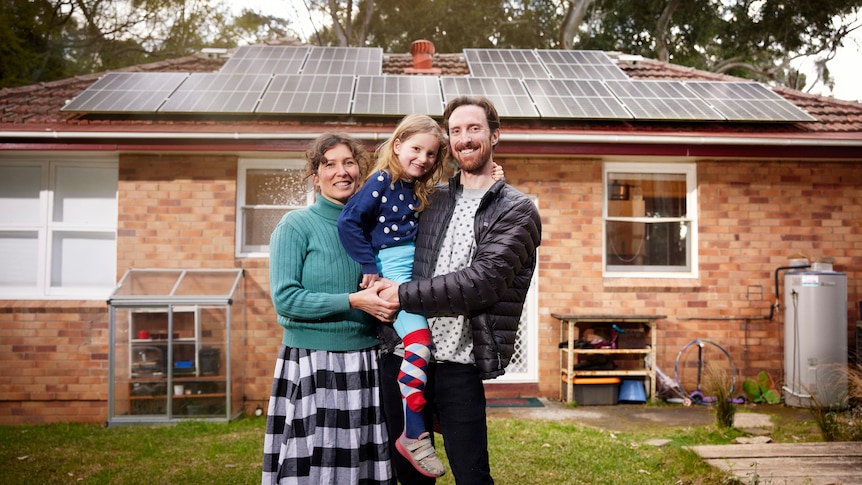A man and woman holding a child stand in front of a modest home with solar panel roof.