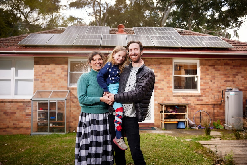 A man and woman holding a child stand in front of a modest home with solar panel roof.