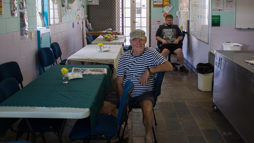 Two men, one in the foreground and one in the background, sit in a cafe with three long tables beside them.