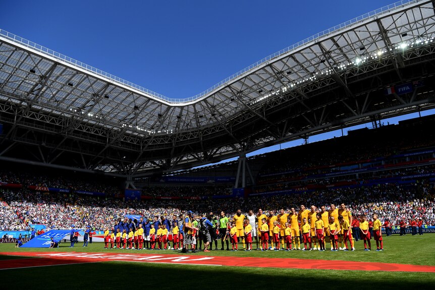 Two soccer teams, one wearing blue and one wearing yellow, line up in a stadium before a game
