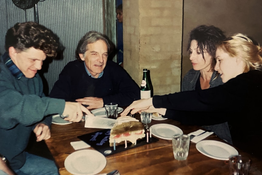 Man and woman their adult son and daughter sit at a table, the son cuts a cake while the daughter serves