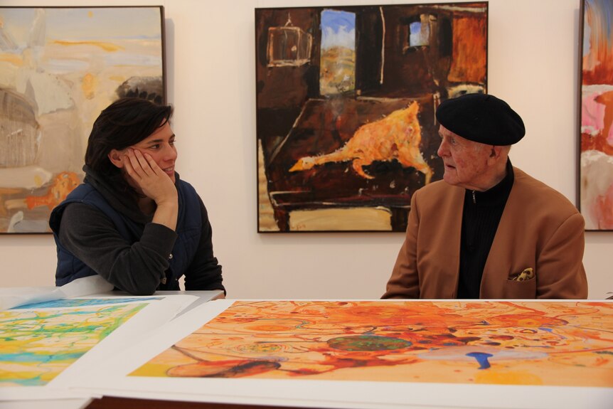 A young women with her chin on her hand sits with an old man in a black beret in a gallery surrounded by paintings