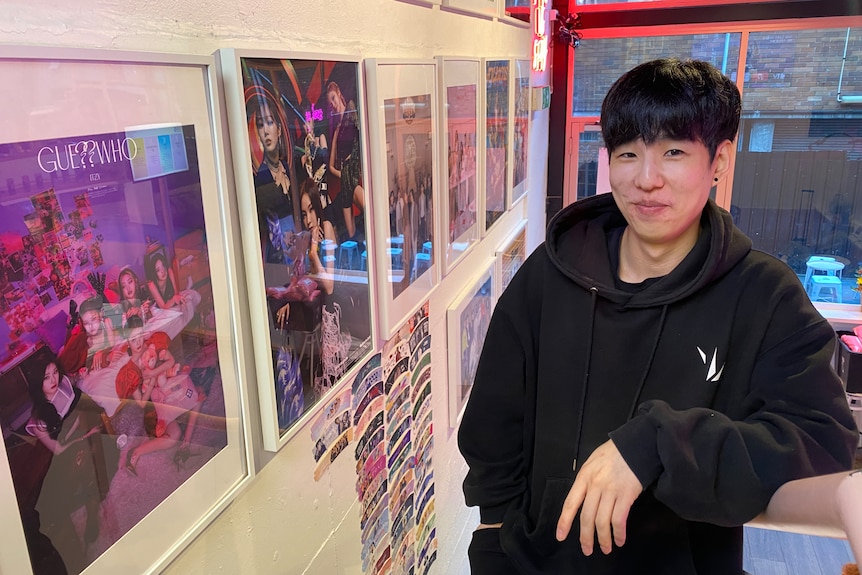 Asian man wearing black hoodie stands next to a wall of posters featuring K-pop artists.