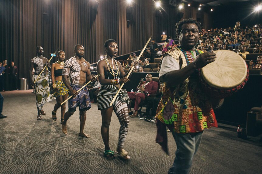 An African drummer in a movie theatre being followed by a group of dancers