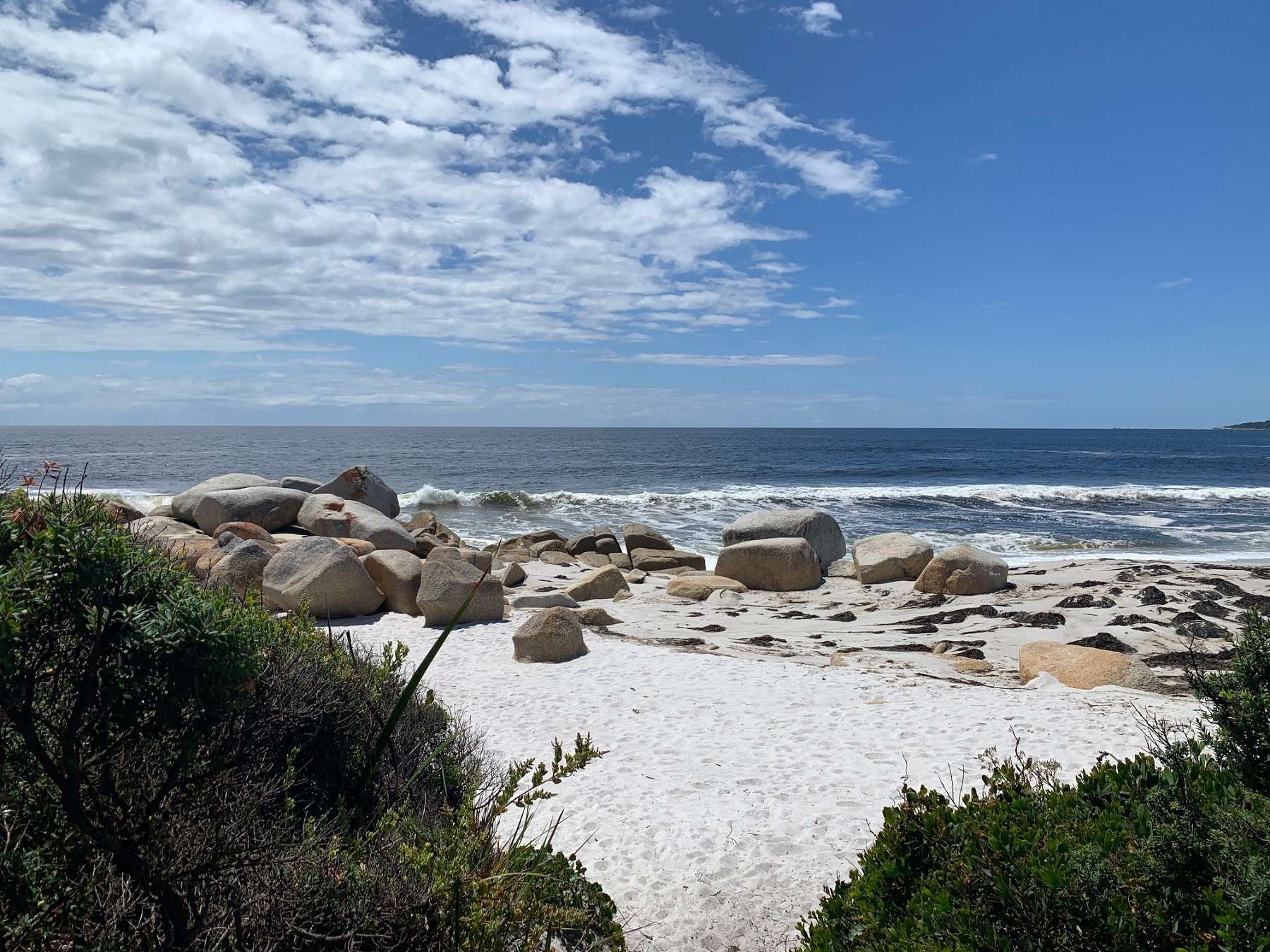 A photo of a beach with white sand and blue ocean with large boulders near the shore.
