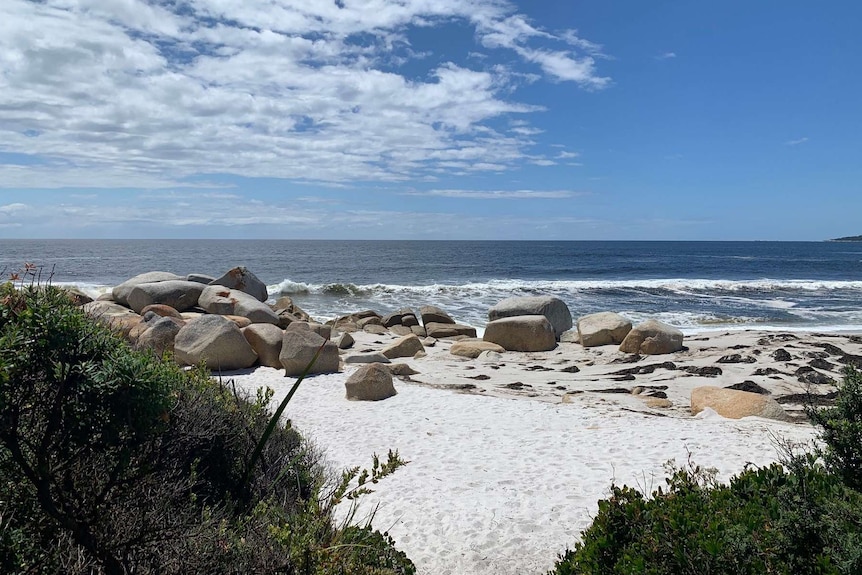 A photo of a beach with white sand and blue ocean with large boulders near the shore.