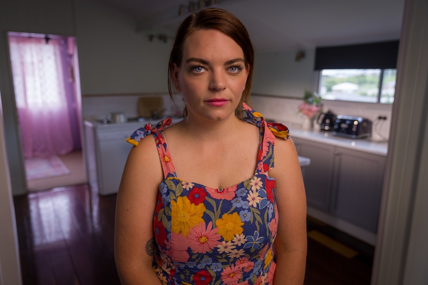 A woman in a colourful dress looks directly at the camera inside a room