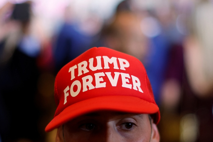 A close up shows a red cap with white text in all caps saying TRUMP FOREVER