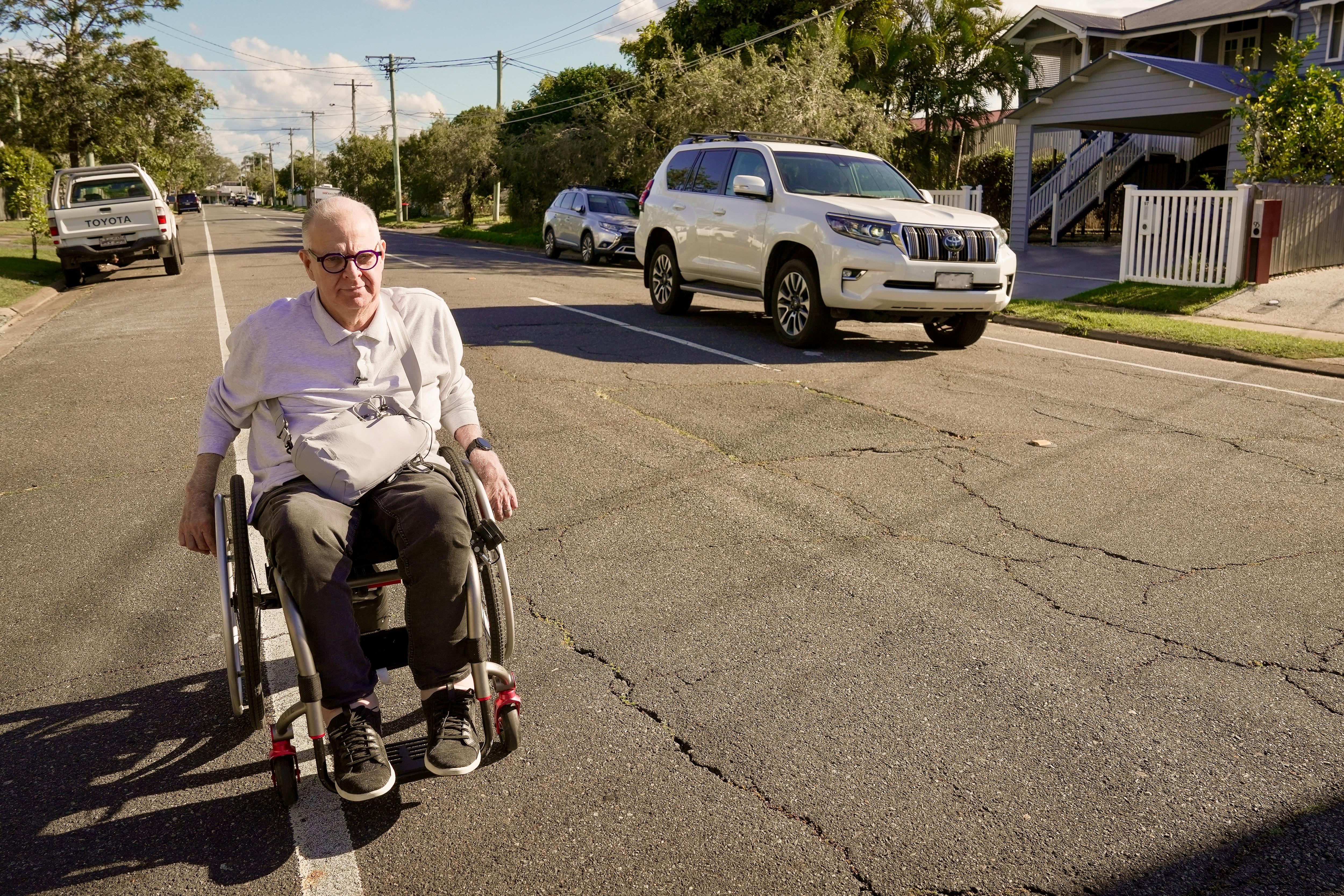 A middle aged white man with short grey hair. He's in a wheelchair, rolling down a surbuban road