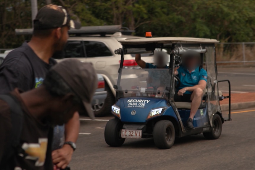 Two men in blue shirts ride on a golf buggy. In the foreground two men stand and look at them.