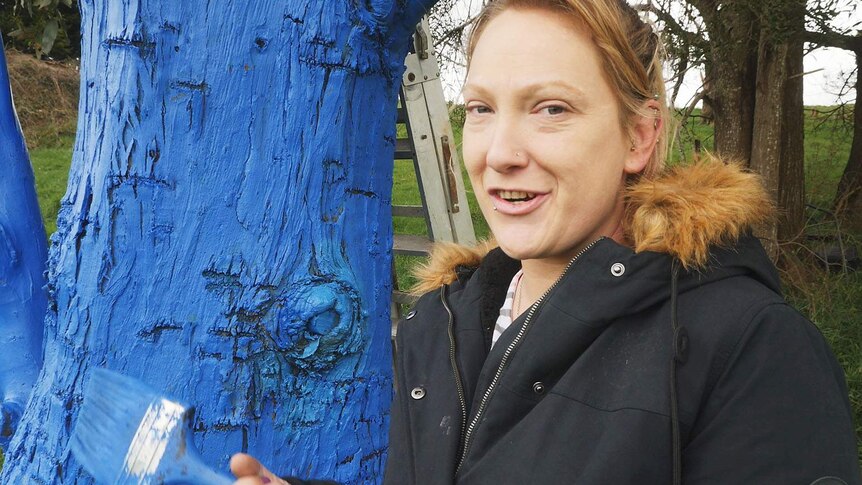 Ash Garth standing next to her blue tree, holding a paintbrush
