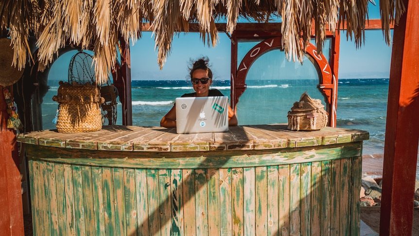 A woman sits at a bamboo desk by the beach with her laptop