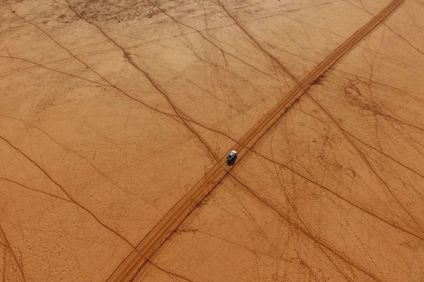 A drone shot of a white ute travelling on a red dirt road, surrounded by the bare outback.