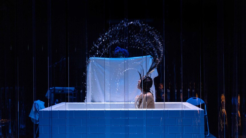 A naked women dramatically flicks her hair out of a bath, a towel in the background, all on stage