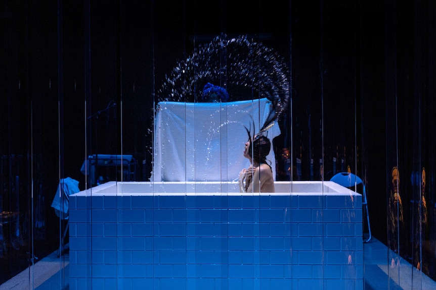 A naked women dramatically flicks her hair out of a bath, a towel in the background, all on stage