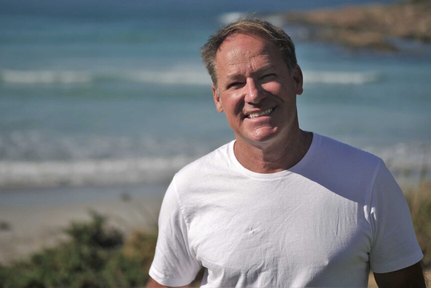 a man in a t-shirt stands and smiles in front of an ocean backdrop.