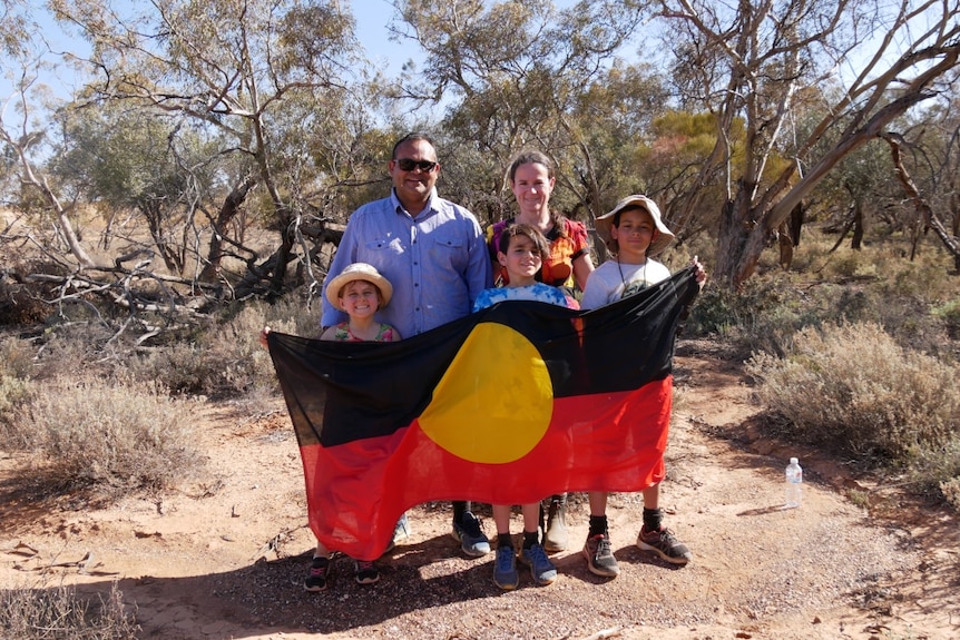 A man, a woman and three children smile at the camera, the children hold and Aboriginal flag.