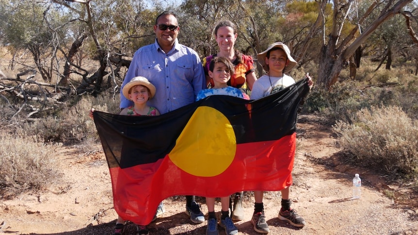 A man, a woman and three childern smile at the camera, the children hold and Aboriginal flag.