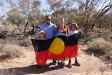 A man, a woman and three children smile at the camera, the children hold and Aboriginal flag.