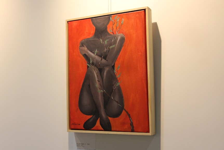 A vibrant red painting hangs on a white gallery wall. The painting depicts a feminine figure cradling herself.