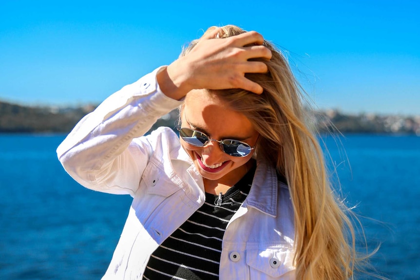 Amy Lyons smiles, looking down, brushing her hair back with the harbour in the background