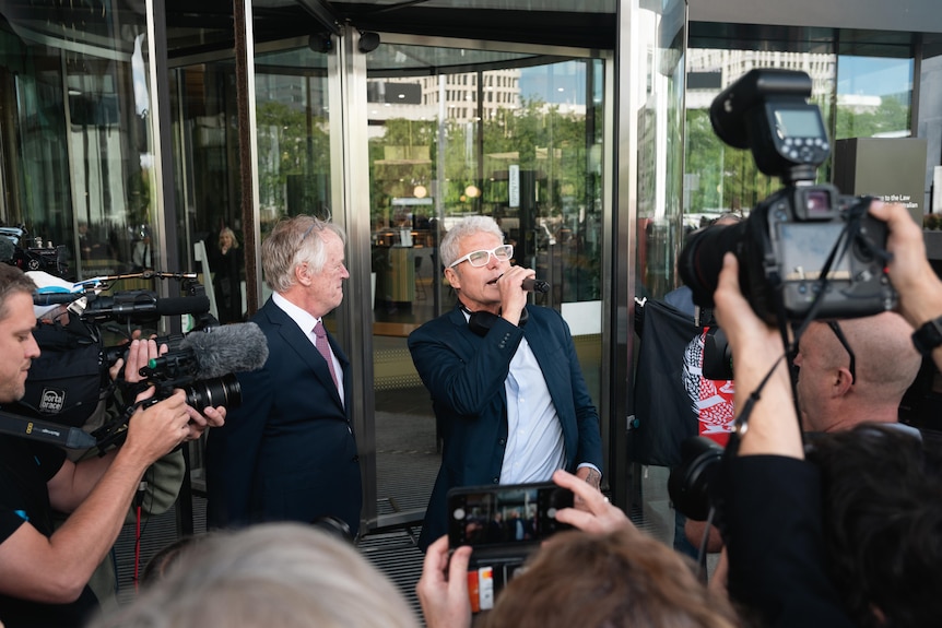 A man standing at a building entrance speaks into a microphone. Around him is a throng of cameras.