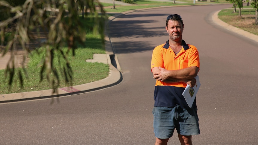 A man in a hi-vis shirt stands with his arms crossed, holding a document, in a suburban street.