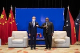 Two men in suits bump elbows as they stand and pose for an official photo in front of the flags of China and PNG.