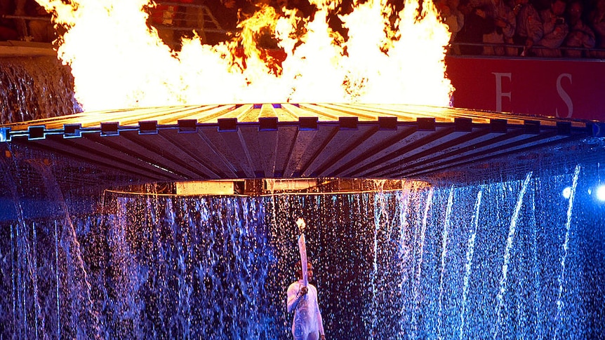 Cathy Freeman stands under a UFO shaped waterfountain, water spilling around her. Above it the Olympic cauldron blazes.