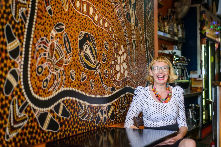 Debbie Guest sits at a table with Aboriginal painting on surface