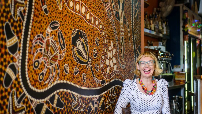 Debbie Guest sits at a table with Aboriginal painting on surface