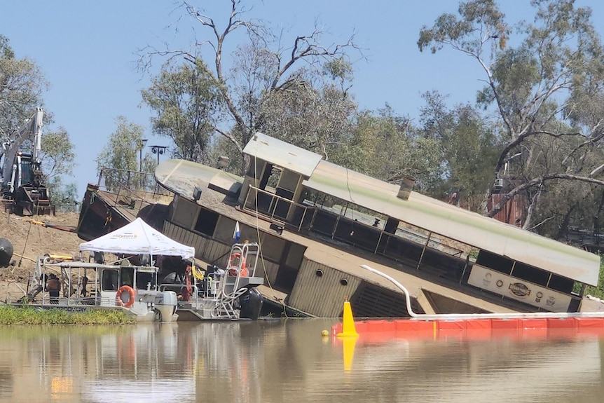 An historic paddle wheeler emerges from the brown water of the Thomson River, with salvage crews on pontoons and machinery.