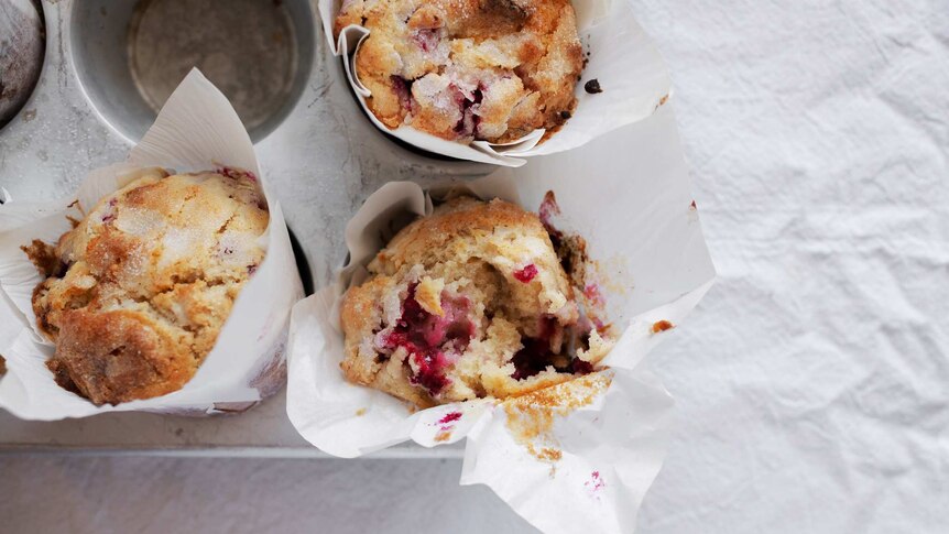 A muffin tray with three freshly baked muffins sitting inside, with raspberry, white chocolate and topped with sugar.