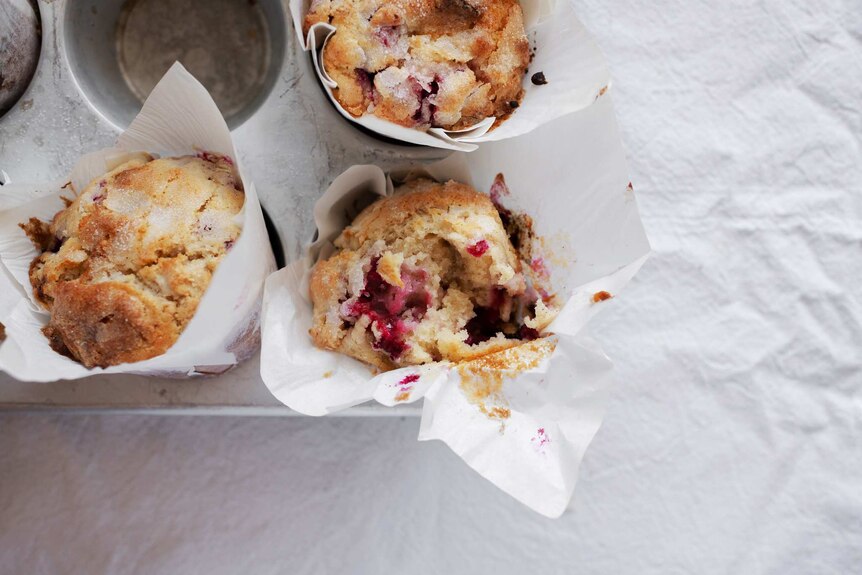 A muffin tray with three freshly baked muffins sitting inside, with raspberry, white chocolate and topped with sugar.