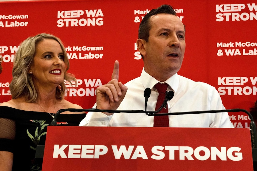 A man in a shirt and tie makes a number one sign during victory speech while standing next to his wife.