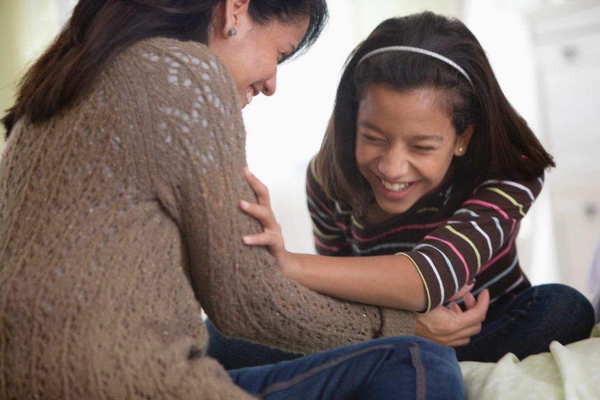 A mother and daughter laugh together while sitting on a bed.