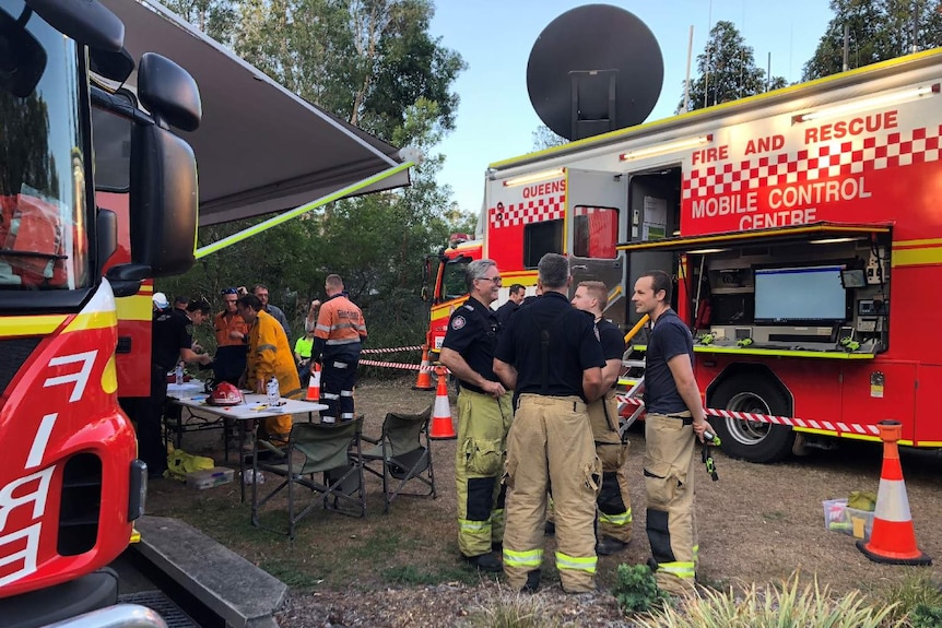 Two fire truck and a half a dozen fire fighters chat in groups.