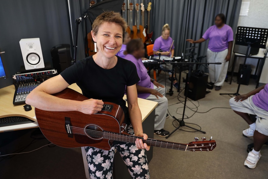 woman sits with guitar smiling with others are recording vocals in a studio behind her 