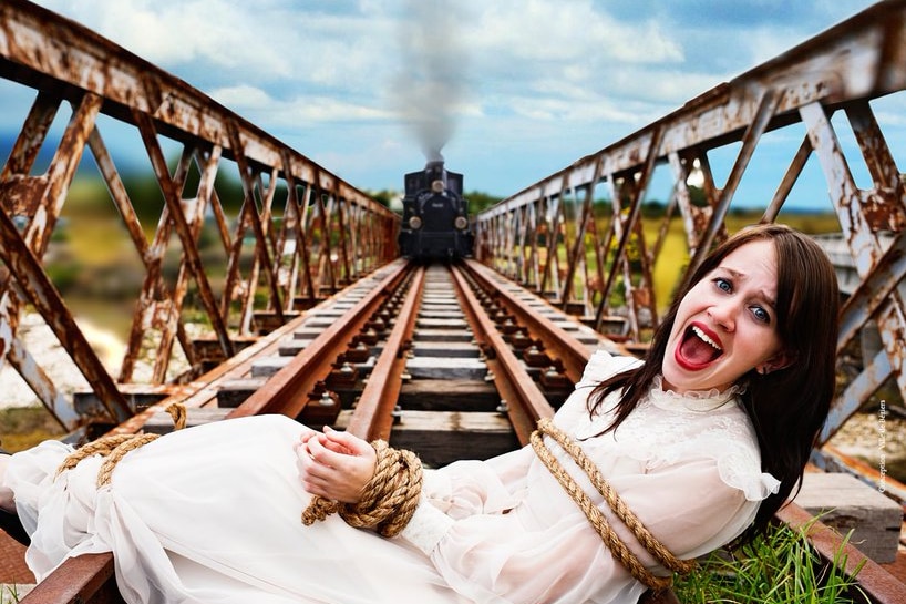 A woman wearing a white dress screams while tied to train tracks with her hands bound as high speed train approaches.