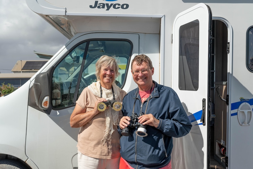 A woman and man holding binoculars stand in front of a campervan.