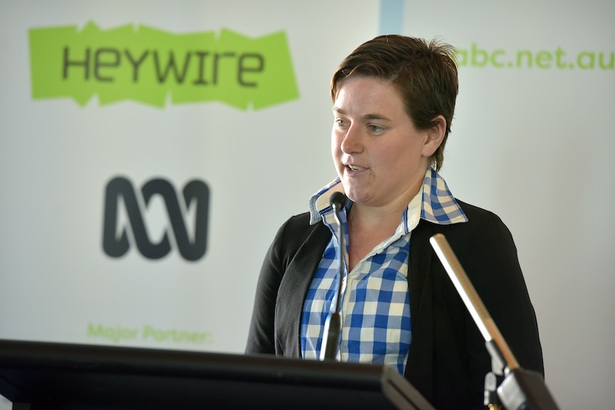 A young woman in a blue check shirt and black blazer stands at a podium with a 'Heywire' sign in the background