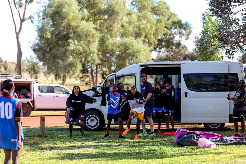 A group of young Aboriginal women look animatedly upward toward an incoming football amid a campground setting with a bus behind