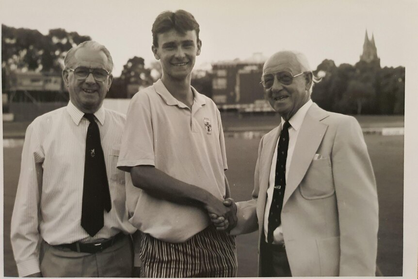 A young boy wearing a white shirt shakes hands with a old man wearing a suit and glasses at a cricket oval. 