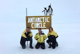 Mark, Peter and Fiona crouch in the snow, in front of a sign that says Antarctic Circle with pictures of penguins on top.