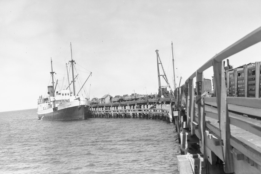 A black and white photo of a fishing boat at the end of a long jetty.